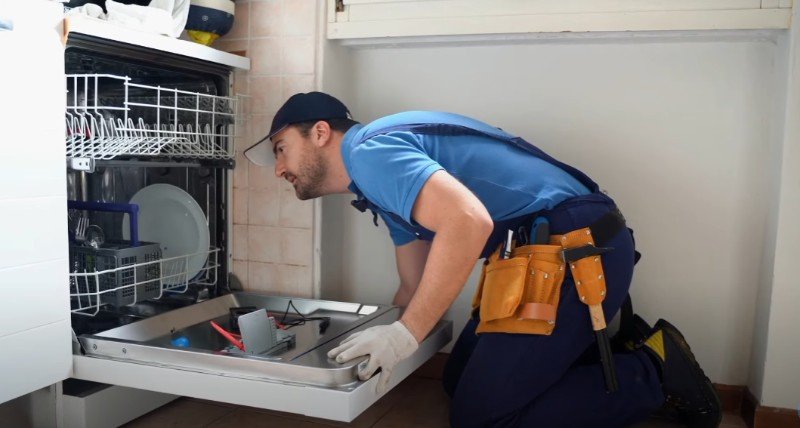 A man in blue overalls repairs a dishwasher in a Pensacola home.