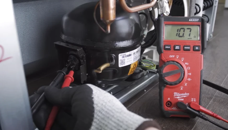 Gloved hand testing refrigerator compressor with multimeter reading 107