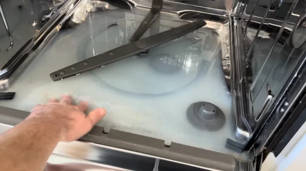 Hand touches soapy water inside open stainless steel dishwasher with spray arm.