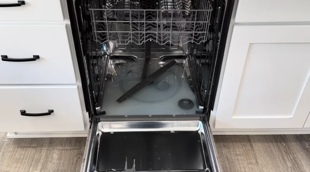 Open dishwasher with racks and wash arm in modern kitchen with white cabinets.