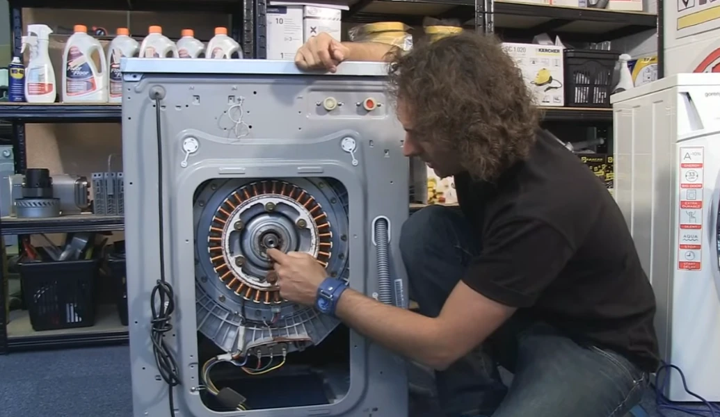 Person examining washing machine repair motor in workshop with tools and detergent shelves