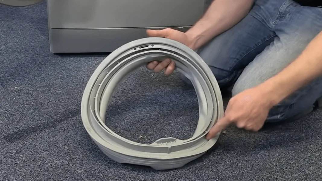 Person inspecting gray washing machine gasket on blue carpet by appliance